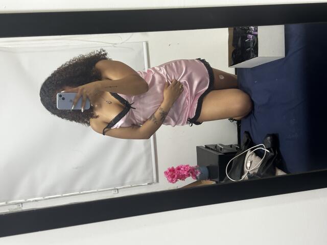 SofiaPassionne with curly hair, wearing a pink satin dress, black bra, and black shorts, kneeling, showing her tits, holding a phone in a mirror selfie.