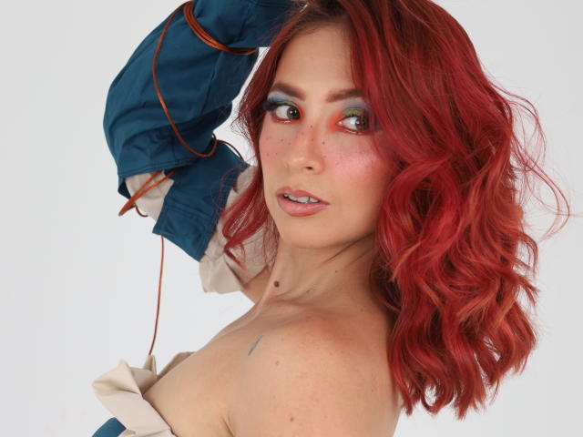 MiaaRouge with red wavy hair, wearing a blue jacket, exposing her tits, green and orange eye makeup, freckles, and a small tattoo on her shoulder. Her expression is sultry.