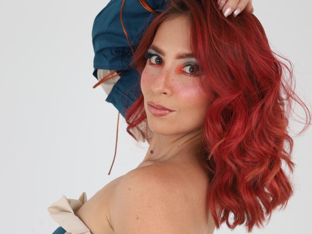 MiaaRouge, met rood golvend haar, groene ogen en sproeten, kijkt over haar schouder, ze draagt een blauw jasje, waarbij haar blote schouder en kleine borsten zichtbaar zijn. Ze glimlacht subtiel..