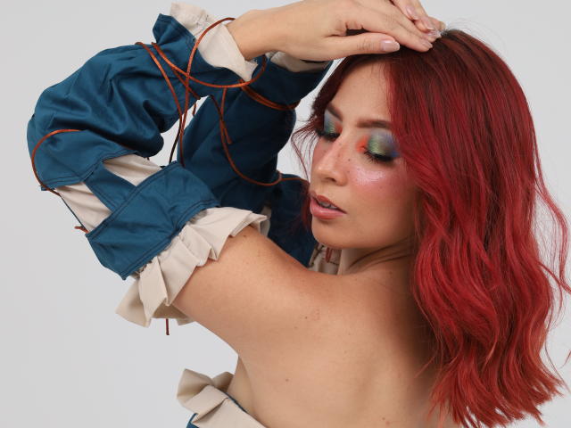 MiaaRouge with red hair, green eye makeup, and a blue jacket with ruffled sleeves, arms raised, showing bare shoulders and a soft, pink lip.