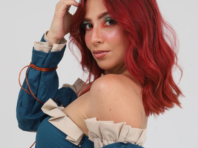 MiaaRouge met rood, golvend haar, draagt een blauwe top met een open schouder en beige ruches, kijkt over haar schouder, met haar hand in haar haar, groene oogschaduw, roze blush en subtiele make-up..