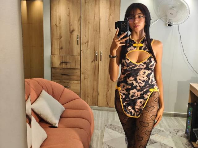 LunaRoja69 in a Chinese-style dress with yellow trim, cut-out boobs, and black fishnet leggings, standing in a room, holding a phone, with a fan in the background.
