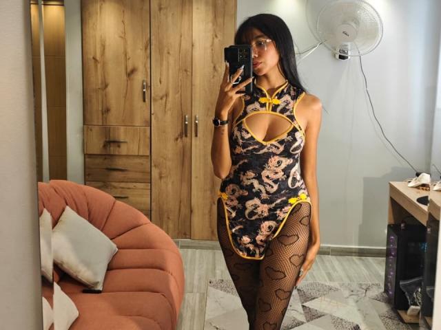 LunaRoja69 in a black floral cheong suit with yellow trim, cutout, and heart-patterned fishnet legs, holding a phone, standing in a room with wooden cabinets.