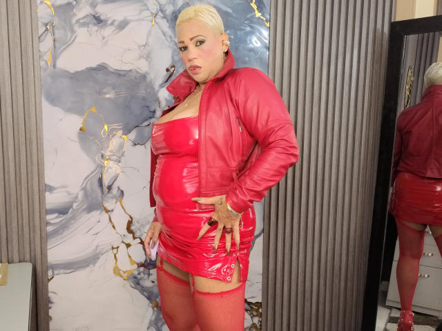 Gianella Cox, con i capelli biondo platino, indossa un abito e una giacca in lattice rosso, mostrando il suo grande sedere e i suoi seni, e porta calze a rete rosse, mentre è in piedi di fronte a uno specchio..