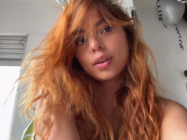 SamantaCarterr with messy, wavy red hair, light skin, and pink lips, looking at the camera with a slight smile, showing a cute, natural face with small nose studs.