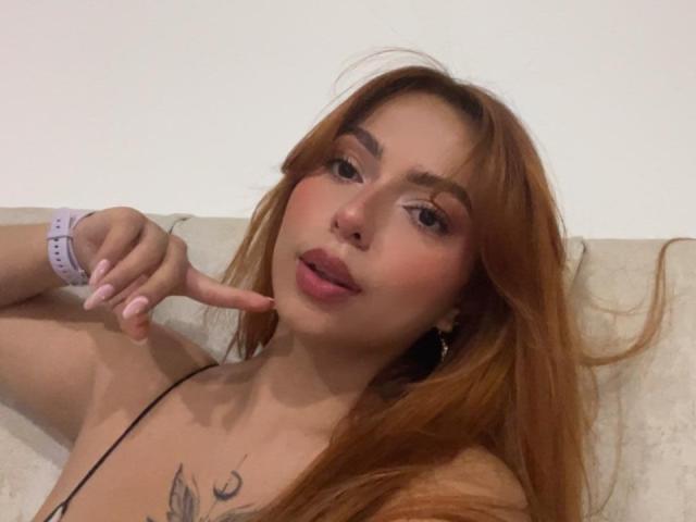 SamantaCarterr with long red hair, pink lipstick, and a nose ring, pointing at her face with a finger, wearing a pink watch, showing tattoos on her chest.