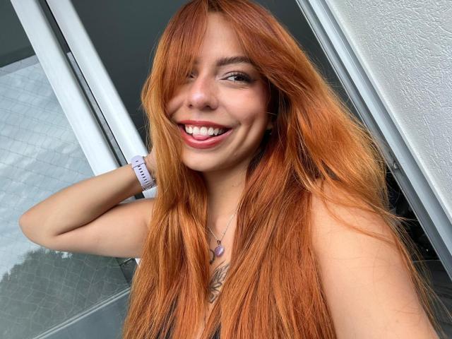 SamantaCarterr with long orange hair, smiling, showing her teeth, wearing a pink watch, hand in hair, tattoo on chest, white wall behind her, looking cute and sexy.