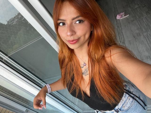 SamantaCarterr with long orange hair, black top, and blue jeans, showing her small tits, smiling, wearing a purple necklace and pink wristband, taking a selfie indoors.