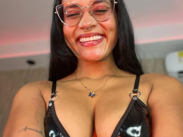LilithRussoo, com óculos, piercing no nariz e um sorriso, vestindo um sutiã preto brilhante que mostra seus seios grandes, um colar com uma pedra azul e tem uma tatuagem no braço direito..