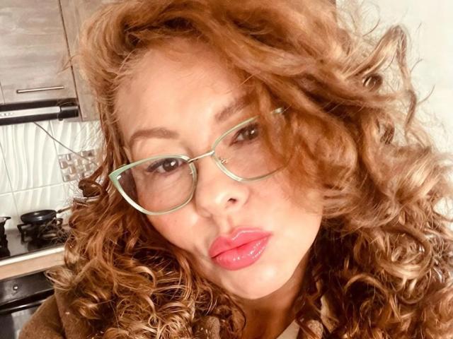 MiaAmoretti with curly brown hair, wearing green glasses, and glossy pink lips, pouting her lips. Boobs not visible, focus on her face. Kitchen background.