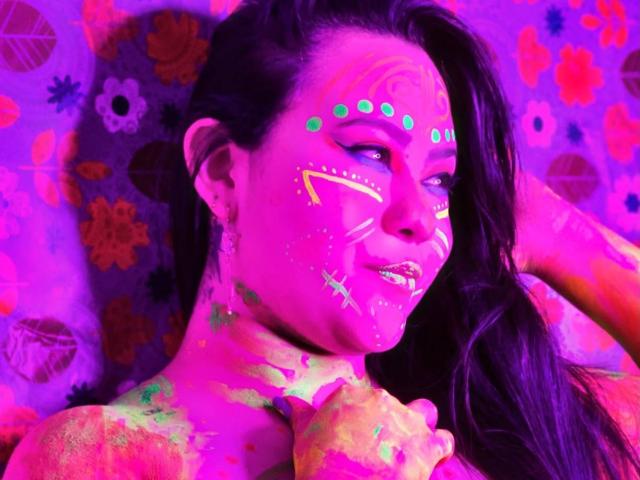 LillyRous has bright pink and purple face paint with green dots, yellow and white lines, and colorful patterns on her tits and neck, smiling with dark hair.