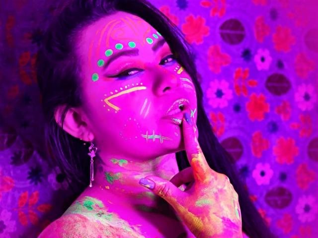 LillyRous has bright pink and green face paint, neon yellow and green patterns, long black hair, and is touching her lips with a finger, wearing a silver earring.