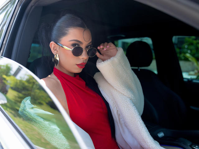 AlissonRossi with dark hair in a ponytail, wearing round sunglasses, a red halter dress, white fuzzy coat, and gold earrings, sitting in a car, adjusting her glasses.