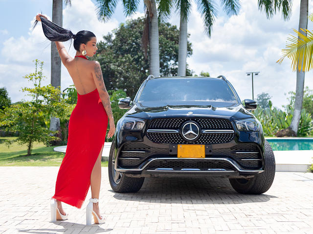 Alisson Rossi, vêtue d'une robe rouge sans manches, porte des talons blancs, tient un haut noir et se tient devant un SUV Mercedes noir brillant. On aperçoit des palmiers en arrière-plan..