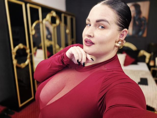 GeorgiaLerox, con i capelli scuri e lisci all'indietro, indossa una maglia rossa trasparente che mette in mostra il suo seno generoso, orecchini dorati, smalto nero per unghie e un'espressione sicura, in una stanza elegante..