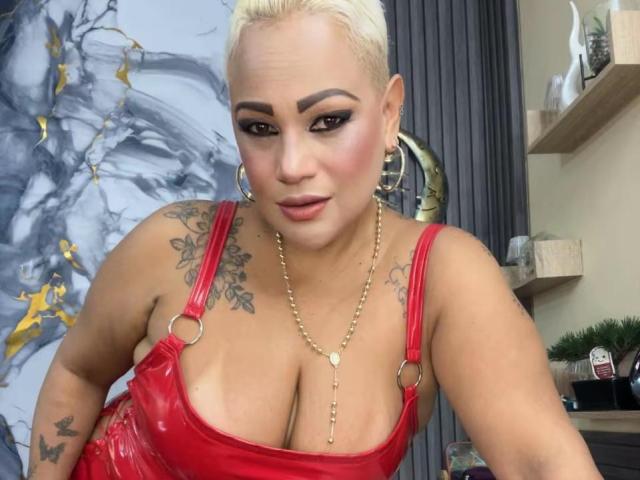GianellaCox with blonde hair, hoop earrings, red vinyl top, gold necklace, tattoos, large boobs, makeup, and a confident expression.