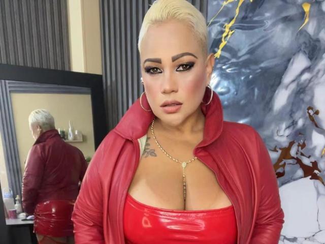 Gianella Cox, con i capelli biondi corti, indossa una giacca e una top in pelle rossa, grandi orecchini a cerchio e una collana d'oro, mostrando il seno in un riflesso allo specchio..