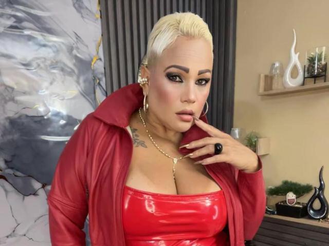 GianellaCox with platinum blonde hair, wearing a red shiny latex top, red jacket, gold necklace, and hoop earrings, touching her chin, showing large tits, and a tattoo on her chest.