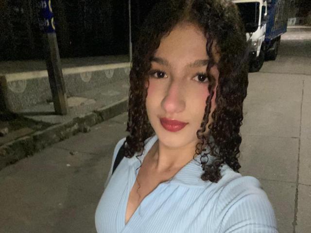 NathachaEvans with curly brown hair, red lipstick, and light blue ribbed shirt, showing her small tits in a nighttime selfie. She's standing on a sidewalk, looking at the camera.