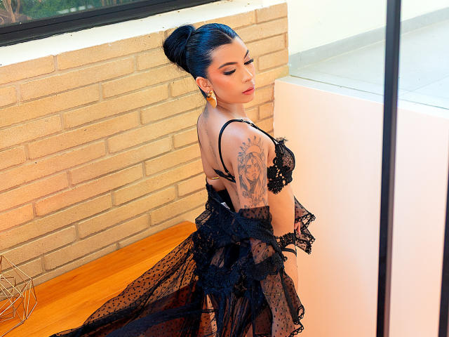 Alisson Rossi con lencería negra de encaje, tatuajes en el brazo derecho, cabello negro recogido en un moño, aretes, apoyada contra una pared de ladrillo, superficie de madera, mirando hacia un lado, vestido negro transparente..