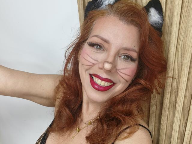Khharha with red curly hair, black cat ears, face paint, and red lipstick, smiling at the camera, showing off her tits and nose, standing against a wooden surface.