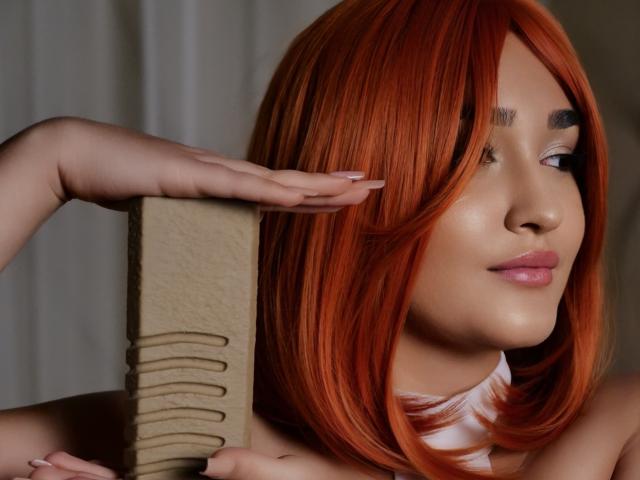 GoldenHear with red hair, holding a beige brush against her head, smiling with pink lips, white necktie, and light skin, showing her ass with her hand in the frame.