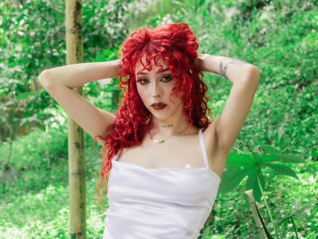 GaiaDoll con cabello rizado rojo, camiseta blanca, brazos levantados, manos en el cabello, tatuaje en el brazo derecho, fondo de bosque verde, usando lápiz labial rojo oscuro, pecho visible, mirando a la cámara..