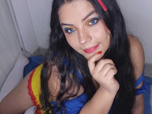 KonyBella with long black hair, blue eye makeup, red lipstick, and a red hair clip, wearing a blue top, yellow skirt, and red lace, resting her chin on her hand.