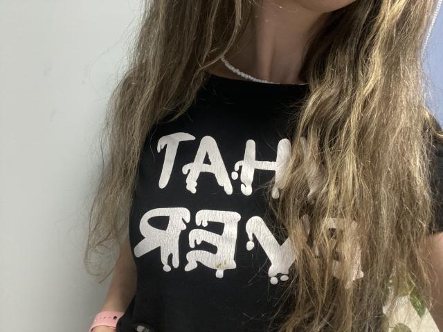 Sanella's chest with long, wavy brown hair, wearing a black shirt with white, drippy text, pink watch, and a white necklace, partially visible face.
