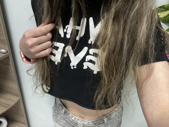 Sanella with long brown hair, black crop top, and grey jeans, holding shirt, showing midriff, pink watch on left wrist.