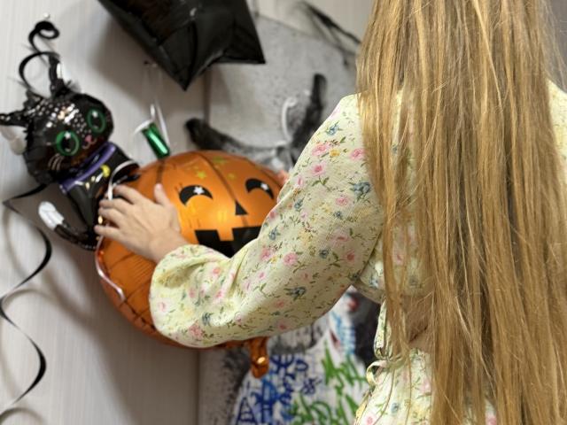 Viloriia with long blonde hair, floral dress, touching a carved jack-o'-lantern, black cat ornament with green eyes, on a white wall. She's decorating, her back to camera.