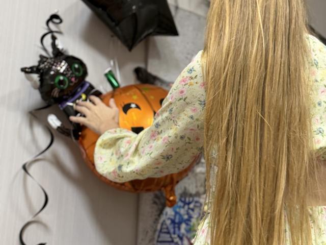 Viloriia with long blonde hair, floral shirt, playing with a black cat mask and orange pumpkin toy, using a green remote control.