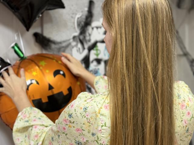 Viloriia with long blonde hair, floral shirt, decorating a large pumpkin with black eyes and a mouth, holding it with both hands.