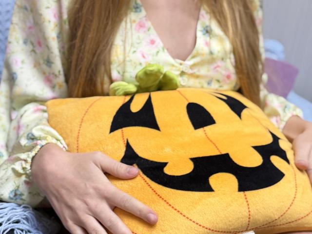 Viloriia with long blonde hair, floral pajama top, holding a yellow Batman pumpkin pillow with black bat emblem, hands gripping pillow.