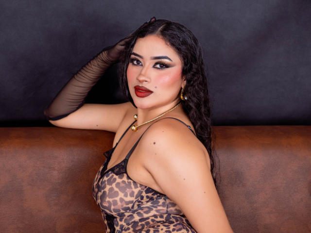AdalinaVera, con lunghi capelli neri, indossa un vestito a stampa leopardo, un guanto di pizzo nero, rossetto rosso e orecchini dorati, seduta su un divano in pelle marrone, che guarda verso la fotocamera..
