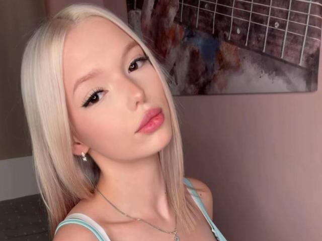 WillodeanAlmonte with blonde hair, light skin, pink lips, subtle makeup, wearing a light blue top, looking at camera, slight tilt, necklace, soft pink wall, abstract art in background.
