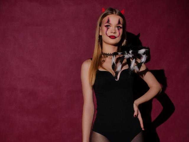MidnightAura with red devil horns, black strapless leotard, black choker, red makeup, black and white feather accessory, hand on hip, standing against red wall.