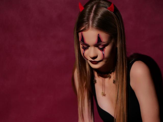 MidnightAura with long brown hair, red devil horns, black makeup, and dark lipstick, leaning forward, wearing a black dress, red background, shadow on the right.