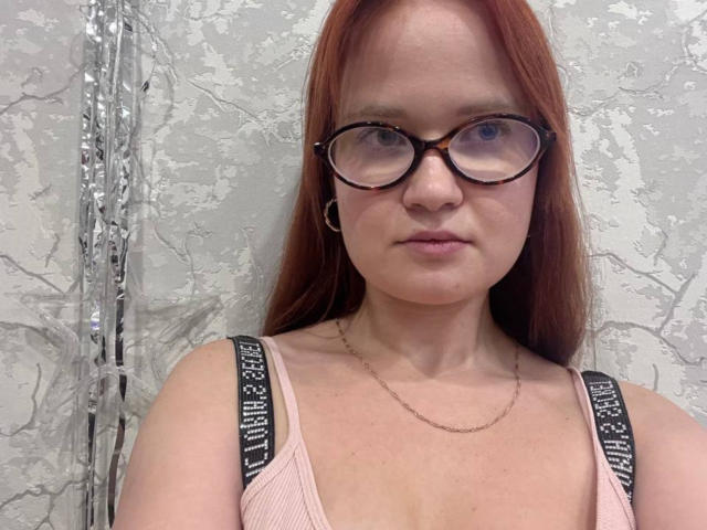 DarleneModest tiene el pelo rojo, gafas y una cara seria. Lleva un top rosa con tirantes negros y un collar delgado. Sus pechos son pequeños y su vulva está oculta..