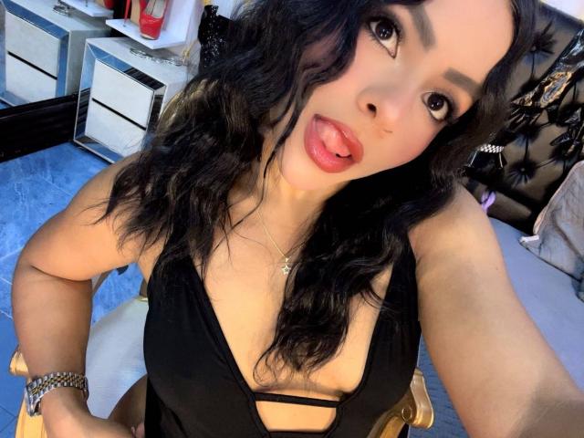 LindaFrancoo, com cabelos pretos ondulados, de língua para fora, vestindo um vestido preto com recortes que revelam seu decote, e um bracelete de prata, em um selfie..