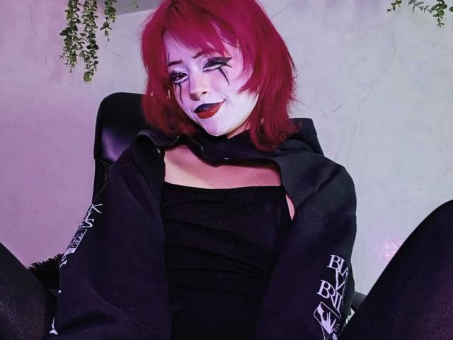 ViolettClay with red hair, white face makeup, black lipstick, black outfit, and black sleeves, sitting with legs spread, smiling, in a chair, wearing a black top.