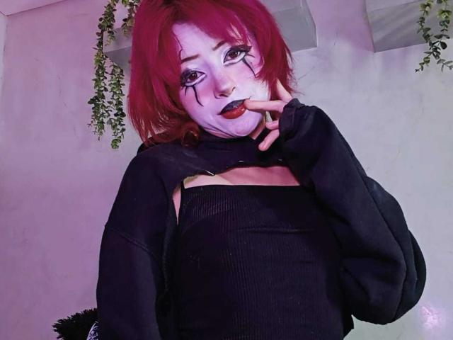 ViolettClay with red hair, black makeup, and black lipstick, finger to mouth, wearing a black cut-out top, black sleeves, in a room with hanging plants.