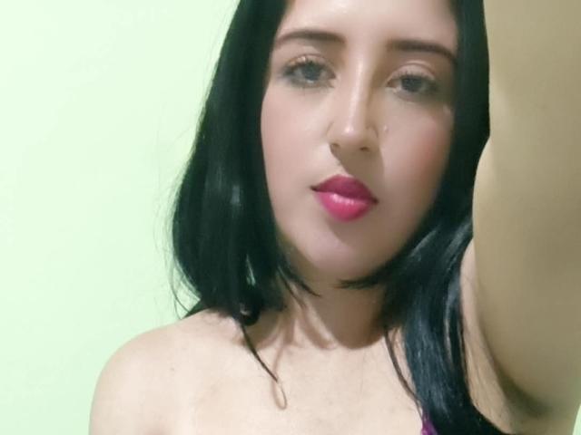 NicollJeans has black hair, pink lipstick, and a nose ring. She's looking at the camera with a slightly raised arm, showing her bare shoulders and upper chest.
