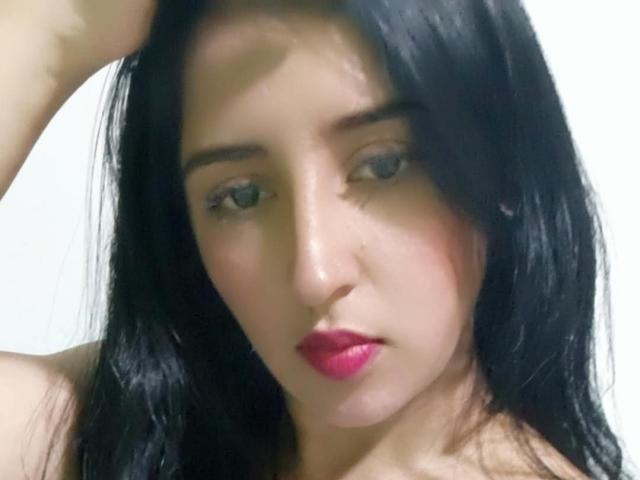 NicollJeans has dark black hair, blue eyes, and pink lipstick. Her right arm is raised, showing a serious expression on her face. She's looking slightly downward.