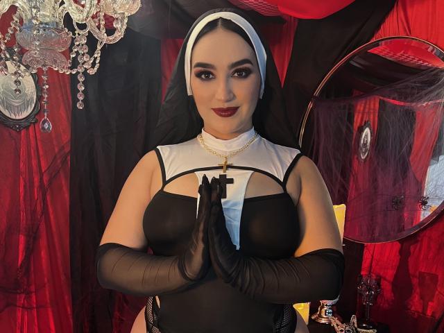 MaraGrief in a black and white nun outfit, with black gloves, holding hands in front, red lipstick, white head covering, cross necklace, and a confident smile.