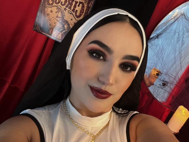 MaraGrief with dark eyebrows, red eyeliner, and deep red lipstick, wearing a nun outfit with a black and white headpiece, gold chain, and white top, smiling at the camera.