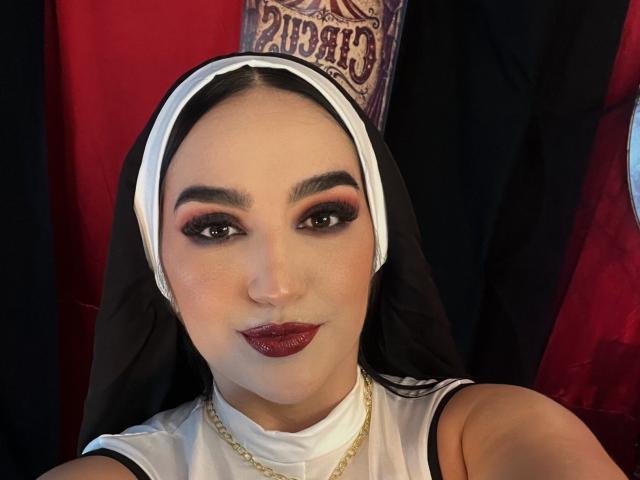 MaraGrief, dressed in a nun's habit, has dark makeup, red lipstick, and a gold chain, staring with sexy, black-lined eyes, and a subtle smile.
