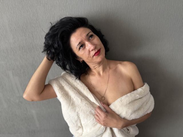 Cameron Enns, com cabelo preto curto, batom vermelho e uma toalha branca e fofa cobrindo seus seios e nádegas, com a mão no cabelo, olhando para cima, com um fundo de parede cinza..