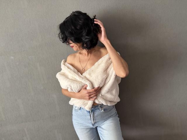 Cameron Enns, com cabelo curto, preto e cacheado, vestindo um suéter branco fofo com um ombro descoberto e jeans azuis claros, com a mão no cabelo, olhando para baixo, usando um colar, magra com seios pequenos, parede cinza..