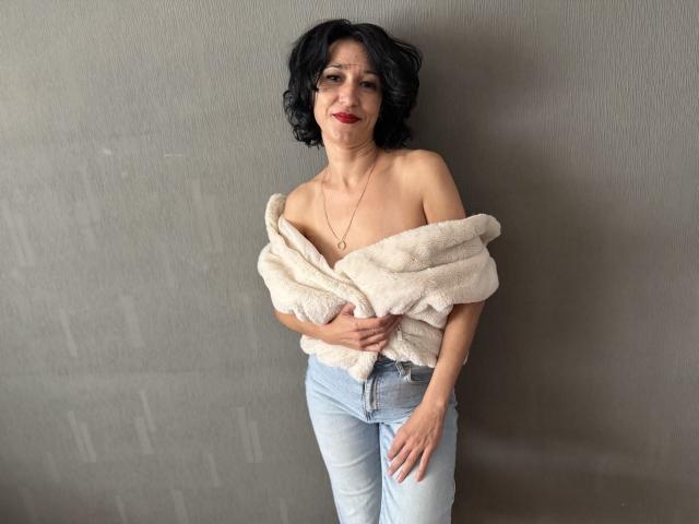 CameronEnns with short black hair, red lipstick, off-shoulder white fleece, and light blue jeans, standing, hands on chest, looking at camera, smiling, necklace visible, gray wall.